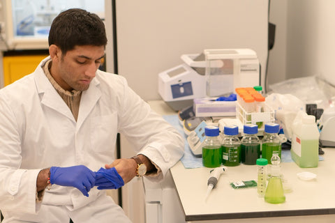 Person in a lab coat and gloves preparing to work with green液体 in a laboratory setting